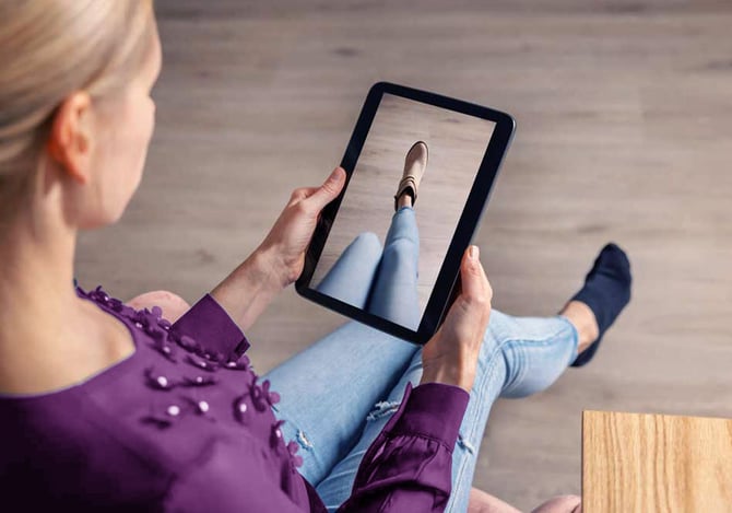 Mercado Pago: Uma mulher com blusa roxa e calça jeans, sentada com um tablet apontado para o pé, tendo uma experiência com a realidade aumentada de um provador virtual.