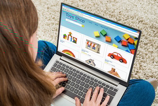 Mercado Pago: menina com cabelos castanhos lisos, vestindo calça jeans, vista de cima, sentada no chão, com o notebook aberto em seu colo em uma loja online de brinquedos, onde ela irá escolher seu presente de Dia das Crianças.