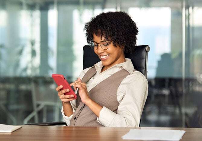 Mercado Pago: Mulher sentada em uma cadeira de escritório, sorrindo enquanto usa um smartphone e organiza o processo de vendas em sua empresa. Ela está vestida com uma camisa clara e colete, com óculos, em um ambiente de trabalho moderno e iluminado.