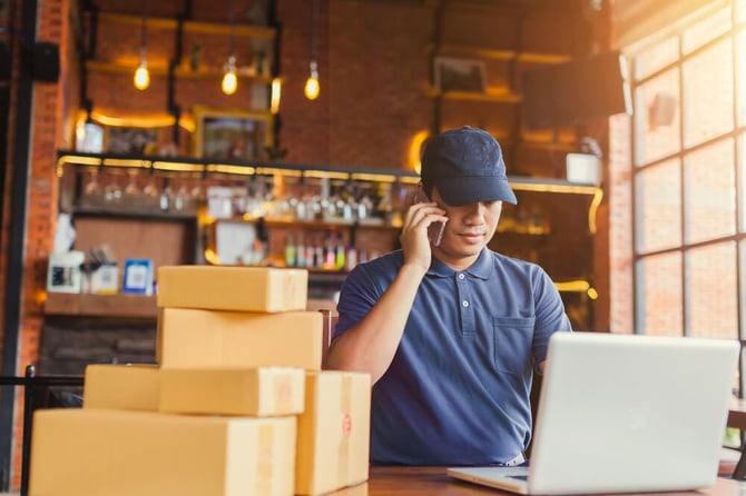 Mercado Pago: Homem com blusa azul e boné falando no telefone olhando para notebook realizando gestão do e-commerce. Compras e pacotes à frente