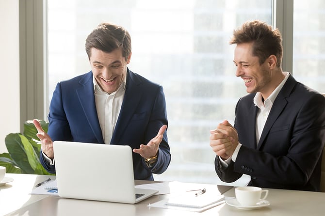 Dois homens vestindo blazer e camisa branca sentados em uma sala de reunião enquanto sorriem para a tela de um laptop devido a análise da taxas de aprovação no e-commerce com auxílio das ferramentas do Mercado Pago