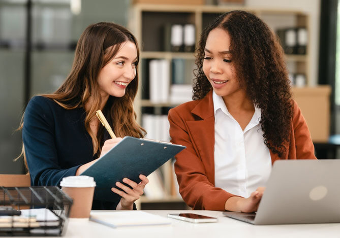 Mercado Pago: imagem de uma empresária acompanhada de uma auditora fiscal. As duas estão sentadas em frente a um notebook, que está sendo usado pela empresária, enquanto a auditora segura uma prancheta e faz anotações para elaborar um checklist para planejar uma auditoria fiscal na empresa.