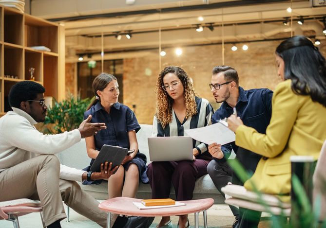 Mercado Pago: Cinco colaboradores, dois homens e três mulheres, sentados em sala de reunião discutindo sobre como tornar o CNPJ da empresa regular. O primeiro homem usa óculos, está vestindo uma blusa branca e calça marrom claro e segura um tablet. A mulher ao seu lado veste um vestido azul escuro e está ao lado de uma mulher de óculos e vestindo uma camisa listrada de preto e branco e utilizando um notebook. Ao seu lado, há um homem utilizando óculos e vestindo uma camisa preta, segurando um papel. Por último, há uma mulher utilizando uma blusa amarela.