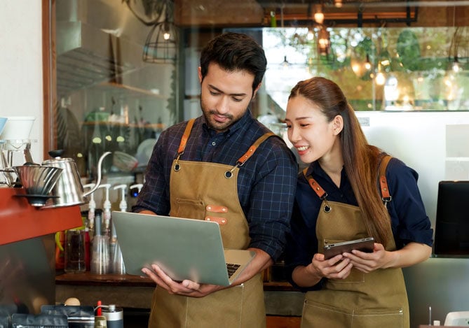segura um notebook aberto e a mulher está com um tablet nas mãos. O homem está apresentando à sua sócia análises que mostram como fazer o parcelamento com máquina de cartão de crédito para que possam escolher a melhor operadora juntos.