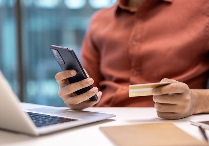 Mercado Pago: imagem de uma pessoa sentada à mesa de trabalho, em frente a um notebook, enquanto segura um celular em uma das mãos e um cartão de crédito na outra, indicando que está fazendo um cadastro de cliente em um clube de assinaturas para cobrança recorrente como uma forma de ter mais previsibilidade de receita e também aumentar o ticket médio da empresa.
