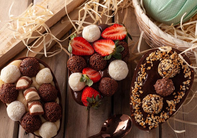 Mercado Pago: imagem de três ovos de Páscoa de colher recheados. Um com brigadeiros e beijos e bombom; outro com beijinhos, brigadeiros e morangos, e o terceiro com pedaços de avelã e três bombons, dois deles cobertos com pedaços de avelã. Os ovos estão sobre uma mesa de madeira, ao lado de uma pequena cesta de vime, onde é possível ver que há um ovo de Páscoa já embrulhado, mas que não aparece por completo na imagem.