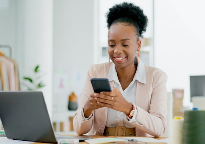 Mercado Pago: Empresária sentada em uma mesa segurando celular e vestindo um paletó rosa claro e uma camisa branca, pesquisando sobre empréstimo empresarial para sua loja.