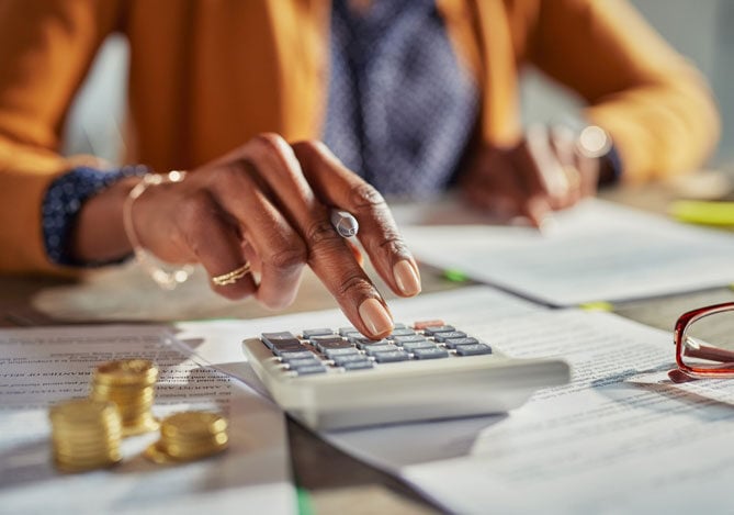 Mercado Pago: imagem das mãos de uma pessoa fazendo contas em uma mesa de trabalho. Em uma das mãos, que está em primeiro plano na imagem, a pessoa realiza cálculos usando uma calculadora enquanto segura uma caneta entre os dedos. A outra mão, em segundo plano na imagem, aponta com um dos dedos para um contrato de fornecimento de linha de crédito empresarial para expansão. Sobre a mesa é possível ver também moedas e outros documentos para avaliação e aprovação de empréstimo empresarial.