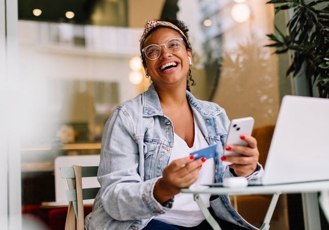 Mercado Pago: Empresária vestindo uma jaqueta jeans, com uma blusa branca e sentada em uma loja, segurando um celular e um cartão e na sua frente, há um notebook branco. Ela está investindo na experiência do cliente unificada da sua empresa. 