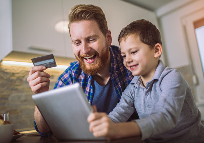 Mercado Pago: Homem sorridente segurando vale-alimentação ou refeição como forma de pagamento, utilizando camisa quadriculada azul e camiseta azul, ao lado de seu filho sorrindo e segurando um tablet, utilizando uma camisa cinza. Ambos estão escolhendo o presente de Dia dos Pais na cozinha de casa. Ao fundo, há armários brancos com luzes. 