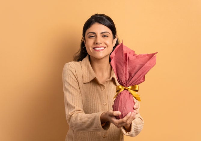 Mercado Pago: imagem de uma mulher sorrindo e segurando um ovo de Páscoa embrulhado em papel na cor coral, com laço dourado, ilustrando uma compra que teria sido feita graças à estratégia de de vendas híbridas que permitiu que a mulher pudesse escolher entre mais formas de pagamento para adquirir seu ovo de Páscoa.