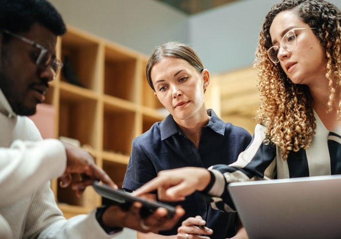 Mercado Pago: imagem de uma equipe reunida em torno de um computador enquanto realizam uma gestão de crise e determinam os melhores passos para colocar em prática um plano de ação que melhore a situação financeira da empresa. Na imagem, é possível ver um homem segurando um tablet com o qual mostra algo aos colegas de equipe, enquanto os demais integrantes do time, duas mulheres e um homem, apontam para o tablet com semblante sério e concentrado. Uma das mulheres está também com notebook aberto.