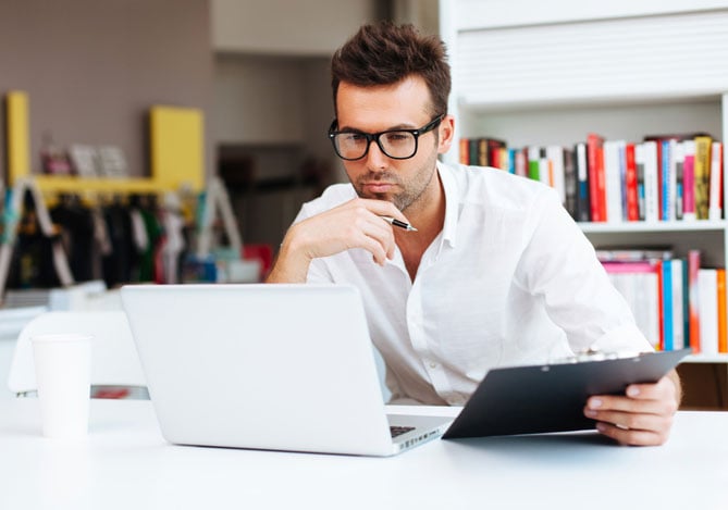 Mercado Pago: imagem de um homem usando uma camisa branca e óculos, segurando uma prancheta, enquanto olha para o notebook ao realizar a declaração de imposto de renda PJ em sua empresa.