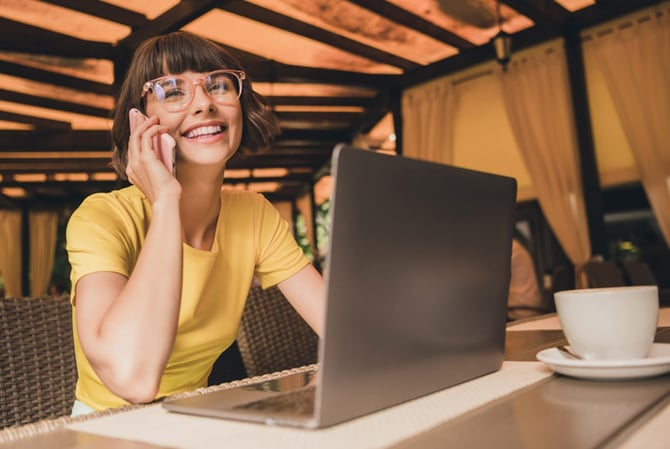 Mercado Pago: mulher usando blusa amarela falando no celular enquanto está com o notebook aberto na mesa com xícara de chá, para reforçar seu trabalho durante a Black Friday e alinhando tudo com a Inteligência Artificial. 