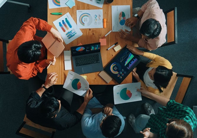 Mercado Pago: imagem de uma equipe sentada em volta de uma mesa de reuniões avaliando relatórios para analisar o market share da sua empresa e, ao mesmo tempo, traçar estratégias de como aumentar o market share. Na imagem, que foi feita de cima, é possível ver seis pessoas, cinco delas sentadas e uma em pé. Todas estão analisando gráficos impressos e também em notebooks e tablets.