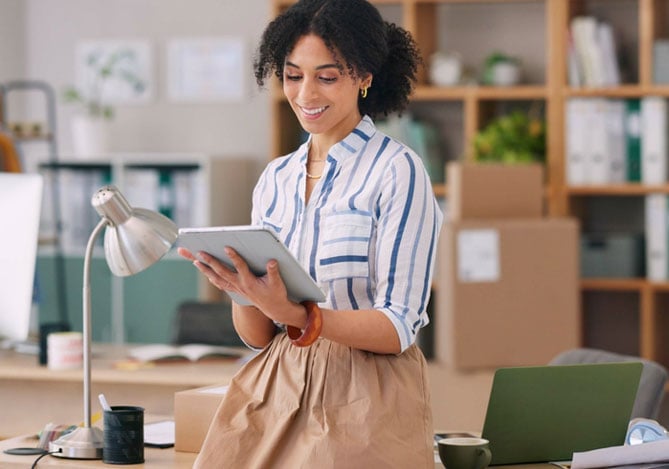 Mercado Pago: mulher sorrindo, com cabelo crespo preso, camisa listrada e saia bege, encostada na mesa do escritório da empresa, analisando os produtos para dropshipping no seu tablet.