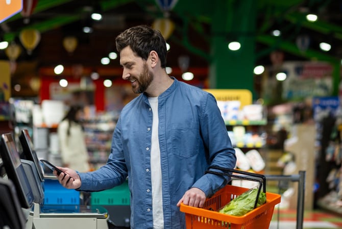 Mercado Pago: homem em mercado, segurando a cesta de compras enquanto realiza o pagamento por aproximação com seu celular no totem de autoatendimento. 