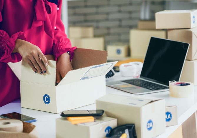 Mercado Pago: Pessoa usando camisa de manga longa vermelha manipulando caixas em cima de uma mesa na qual há celulares e demais objetos, como fitas adesivas, usados para empacotamento de produtos em empresas do mercado digital.