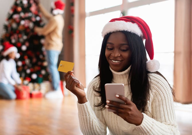 Mulher com gorro do papai noel mexendo no celular enquanto faz compras no Natal digital com ajuda do Mercado Pago