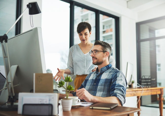 Mercado Pago: imagem de um empresário sentado em frente a um computador em um escritório enquanto faz a busca para a seleção correta da CNAE da sua empresa junto com sua sócia, que está em pé atrás, atrás de sua cadeira e gesticulando com as mãos. Em cima da mesa é possível ver alguns objetos de escritório, como agenda e bloco de notas.