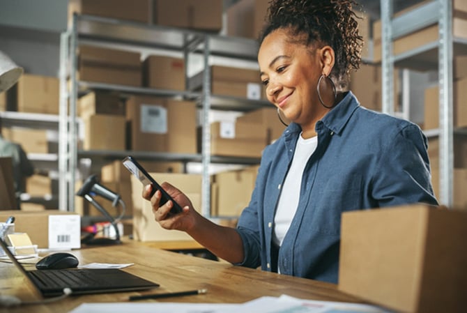 Mercado Pago: mulher em armazém com várias caixas, sentada à mesa, com notebook à sua frente e celular em mãos para realizar o planejamento operacional da sua empresa. 