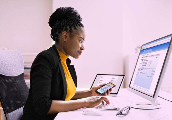 Mercado Pago: mulher com paletó preto e camisa amarela, sentada em cadeira branca e mesa branca, realizando o seu planejamento tributário através do computador e celular em mãos. 