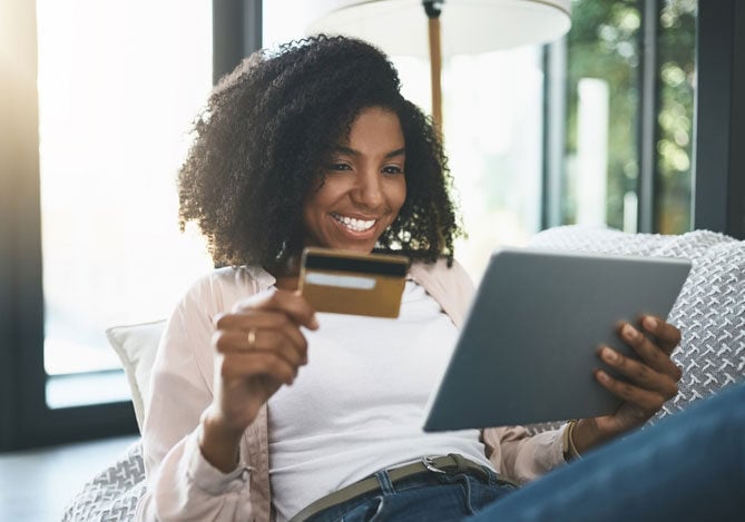 Mercado Pago: Mulher sorridente relaxa no sofá enquanto faz uma compra online utilizando um tablet e um cartão de crédito, representando praticidade e segurança em planos de assinatura.