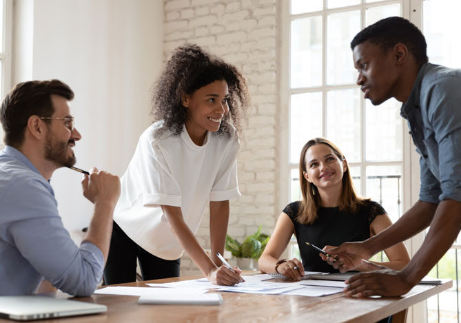 Mercado Pago: imagem de quatro pessoas em volta de uma mesa de reuniões, duas mulheres e dois homens vestindo camisa azul, camiseta branca, camiseta preta e camisa cinza, conversando sobre contratação de força de trabalho enquanto calculam quanto custa um funcionário para a empresa. Sobre a mesa é possível ver alguns documentos com registros e números da empresa.