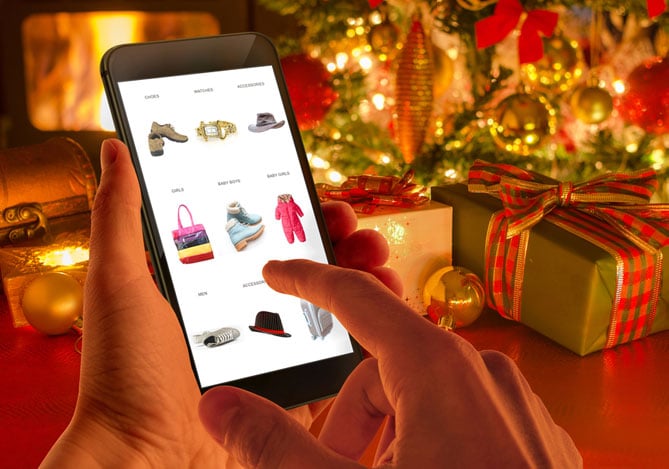 Mercado Pago: imagem das mãos de uma pessoa segurando um celular enquanto faz compras online para o Natal em um site com checkout otimizado para vendas mobile. Na imagem do celular, é possível ver que o site é de roupas e acessórios e, ao fundo da foto, é possível ver decorações de Natal, como árvore e caixas de presente