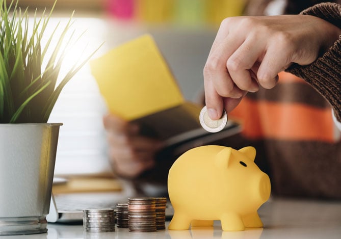 Mão de uma pessoa colocando moeda em um porquinho amarelo, além de outras moedas em uma mesa, sugerindo criar uma reserva financeira e fazer o seu dinheiro automaticamente no Cofrinhos Mercado Pago.