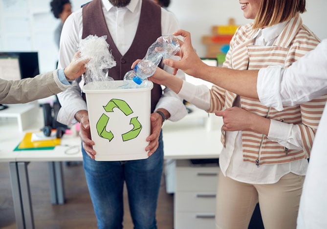 Mercado Pago: colaborador com camisa branca, colete vermelho e calça jeans segurando lixo reciclável para outros três colaboradores jogarem lixo, celebrando a Semana do Meio Ambiente. Ao fundo, há um escritório com mesas. 