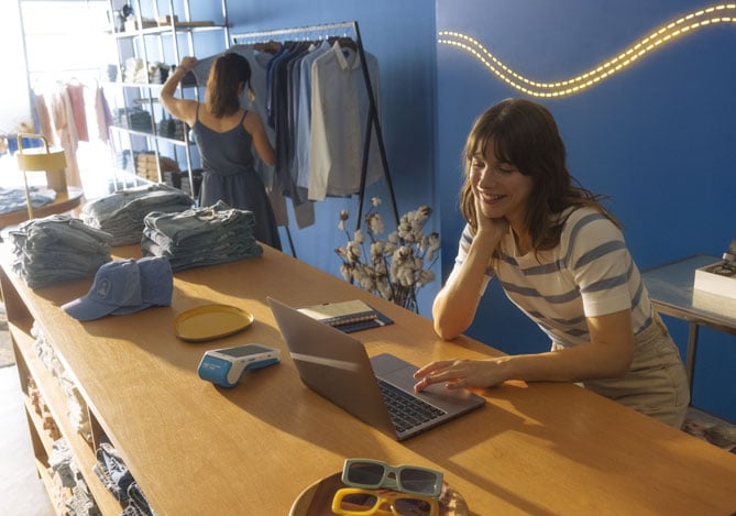 Mercado Pago: Empreendedora sorridente gerenciando seu negócio em um laptop dentro de uma loja de roupas, utilizando um sistema de gestão integrado para otimizar as vendas e organização financeira.