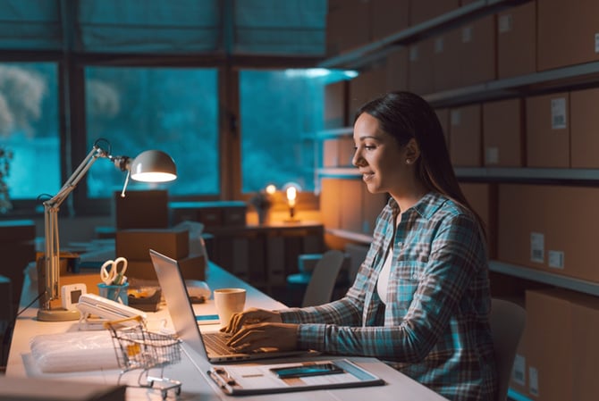 Mercado Pago: empreendedora em seu local de trabalho, onde há uma prateleira com caixas atrás dela, enquanto ela está mexendo no notebook em uma mesa para estudar como aumentar o ticket médio