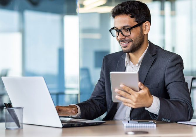 Mercado Pago: imagem de um empresário de óculos sentado em sua mesa de escritório vestindo camisa branca e terno azul, enquanto mexe em um notebook e avalia a eficiência operacional de sua empresa com a ajuda de um tablet.