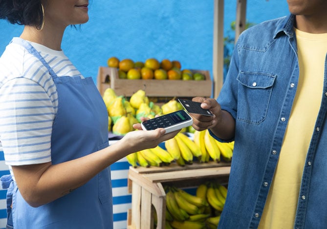 Mercado Pago: vendedora em sua venda de frutas com a máquina de cartão na mão enquanto o cliente que está na sua frente aproxima o cartão Mercado Pago para realizar o pagamento. 