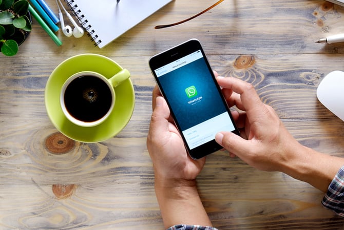 Mercado Pago: Mesa vista de cima, onde estão apoiadas uma xícara de café, cadernos e as mãos de uma pessoa segurando um celular com o símbolo do WhatsApp, representando alguém que vai vender pelo WhatsApp.