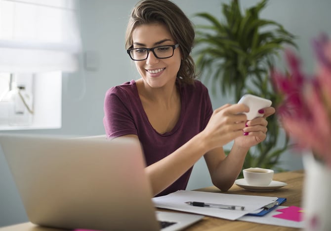 Mulher de óculos sorrindo enquanto segura seu smartphone e vê o laptop