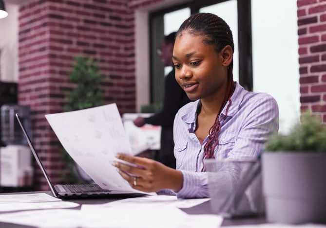 Mulher com blusa roxa trabalhando em gestão financeira no escritório e segurando papel, sentada em mesa de escritório, com destaque para o uso do Mercado Pago como plataforma de pagamento online