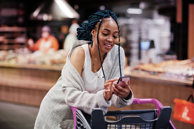 Mulher mexendo em seu celular enquanto faz compras no supermercado com seu cartão Mercado Pago Beneficios