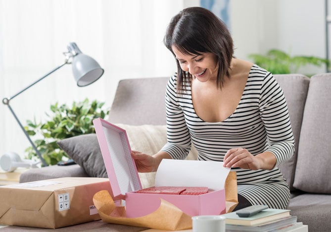 Mercado Pago: Mulher feliz e animada em casa, segurando uma caixa em suas mãos enquanto sorri e fazendo unboxing de um produto