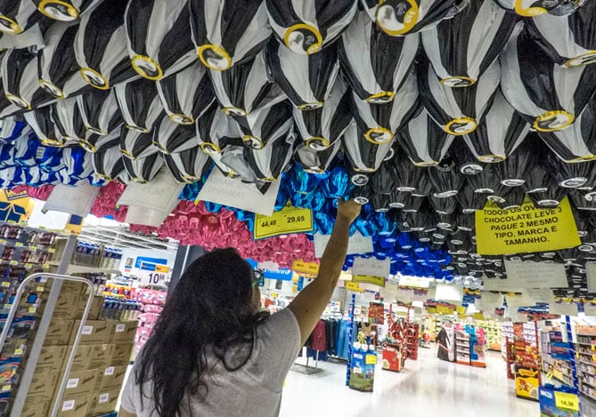 Mercado Pago: Mulher de costas com blusa cinza em supermercado, escolhendo ovos de Páscoa com o vale alimentação. 