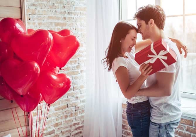 Mercado Pago: Casal com blusa branca, segurando presente vermelho em sala com balões em formato de coração vermelho comemorando o Valentine’s Day