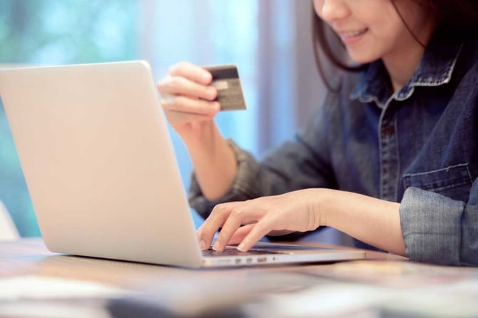 Mulher vestindo uma camisa jeans olhando seu cartão de crédito enquanto digita suas informações em um computador apoiando em uma mesa para finalizar o processo de gateway de pagamento com a ajuda do checkout otimizado do Mercado Pago