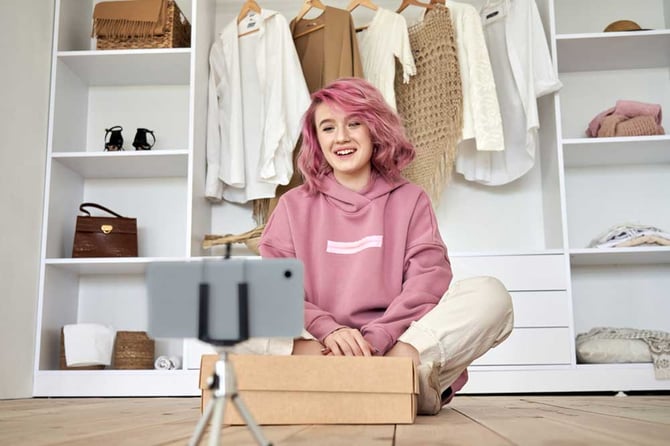 Mercado Pago: Influenciadora digital com moletom rosa sentada em seu closet com celular em tripé e caixa em sua frente,  criando uma estratégia de marketing digital para e-commerce. 