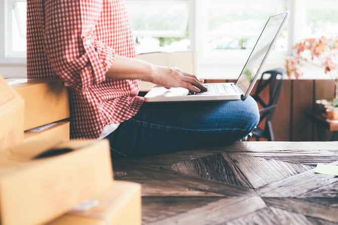 Mulher vestindo calça jeans e camisa quadriculada vermelha e branca sentada no chão com as pernas dobradas, enquanto mexe no laptop apoiada nas suas pernas para verificar mais informações sobre as disputas de contestações em compras online e como o Mercado Pago pode auxiliá-la