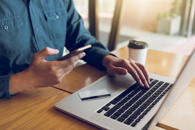 Mãos de um homem mexendo em um laptop e na outra um celular para fazer a integração da carteira digital Mercado Pago com o checkout de pagamento da sua loja online