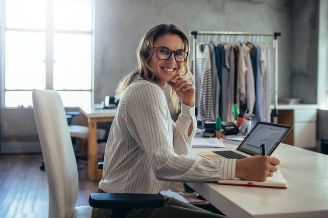 Mulher com camisa listrada , sentada em mesa de escritório com notebook aberto na API Mercado Pago e anotando informações em seu caderno