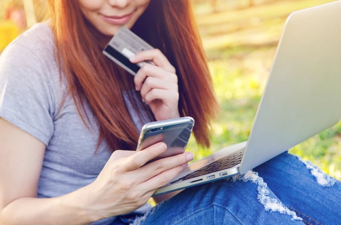 Mulher sentada com blusa cinza e calça jeans, segurando cartão e celular nas mãos e notebook apoiado no colo buscando sobre segurança em pagamentos online.