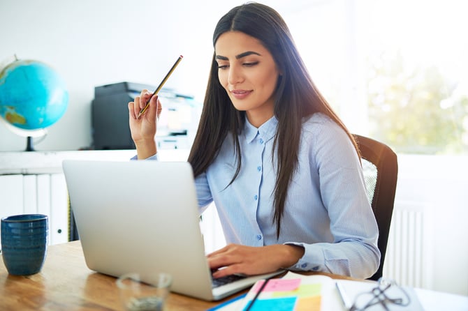 Mercado Pago: Mulher com blusa azul clara sentada em mesa de escritório, com notebook aberto de sua frente com plataforma de e-commerce e segurando lápis em outra mão. 