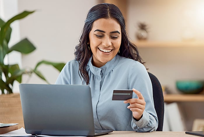 Mercado Pago: mulher sorrindo e sentada à mesa, com o seu notebook e um cartão na mão, representando uma gestora de empresa que está estudando como receber pagamento cartão de crédito sem prejudicar as finanças.